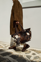 A model walking outdoors holding a dark brown and cream cow print pattern tote bag with dark brown rope handles , wearing a long brown coat and white pants with dark brown shoes.