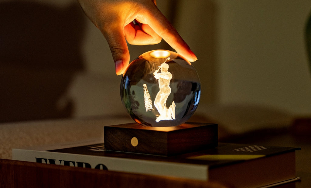 Gingko Design – Amber Crystal Light - Golf, een hand raakt een oplichtende kristallen bol aan die op een boek ligt
