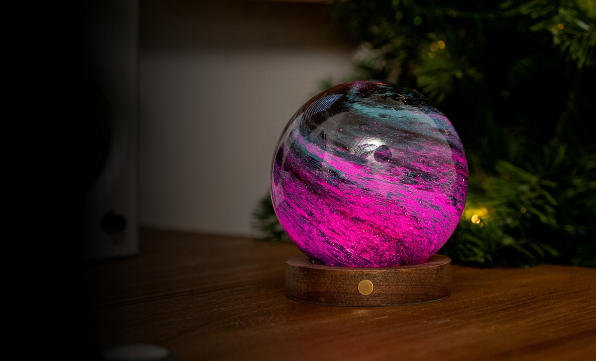 Gingko Design – Leuchte Astraglass - Stellar Ocean - 2 Größen, dekorative leuchtende Kugel in Rosa und Blau auf einem Holzsockel neben einem Tannenbaum