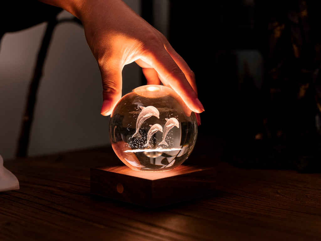 Gingko Design – Amber Crystal Leuchte - Delfine, eine Hand berührt eine leuchtende Glaskugel mit eingravierten Delfinen