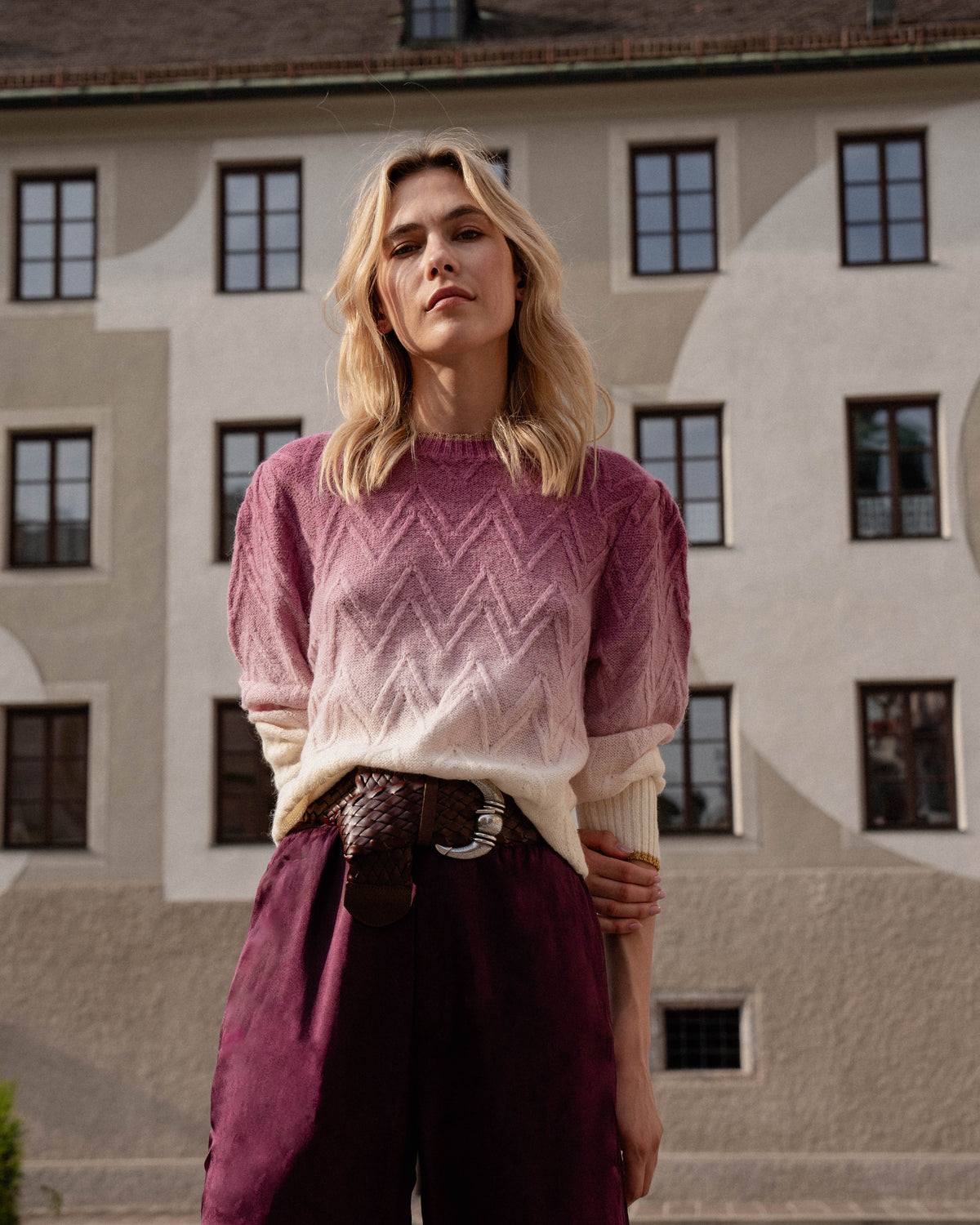 Michonet – Hose Silk Berenjena, blondes Model in rosa-weißem Pullover, bordeauxroter Hose, geflochtener Gürtel