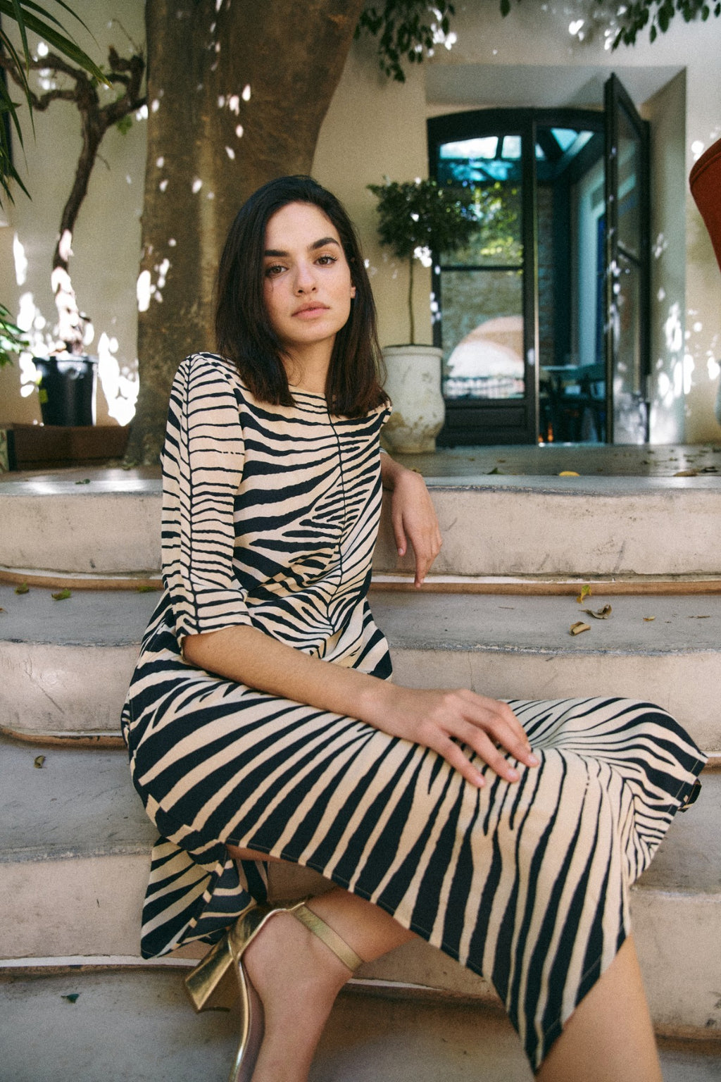 Michonet – Kleid Hoja Beig, sitzendes Model von vorne, Zebrakleid in Beige und Schwarz, goldene Schuhe