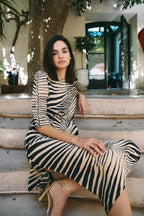 Michonet – Kleid Hoja Beig, sitzendes Model von vorne, Zebrakleid in Beige und Schwarz, goldene Schuhe