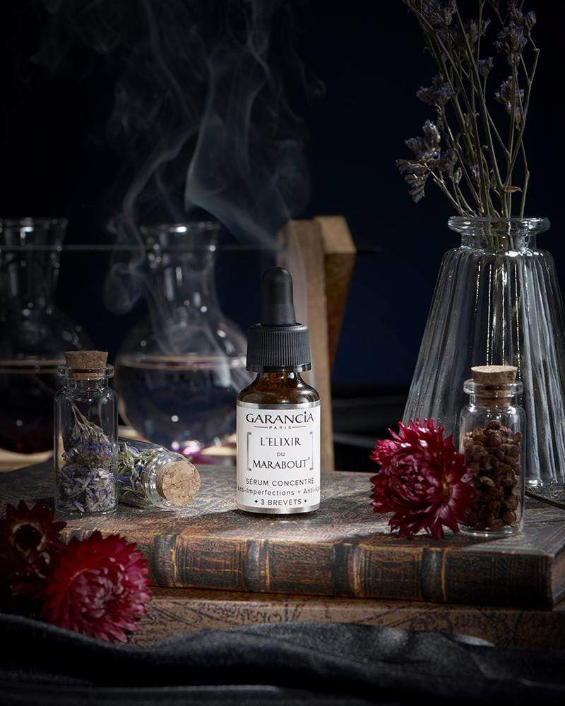 Sérum L'Élixir du Marabout de Garancia posé sur un vieux grimoire, entouré de fleurs rouges, de fioles d'apothicaire et d'une fumée mystique évoquant un laboratoire de sorcellerie.