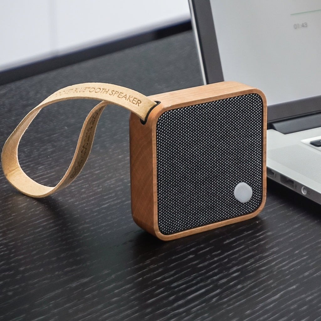 Gingko Design – Bluetooth-luidspreker MI Square - Natuurlijk kersenhout, houten bluetooth-luidspreker op een bureau naast een computer