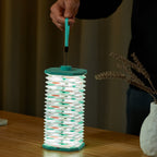 Gingko Design – Lampe Smart Accordion - Velours Bleu Géometrique, une personne tient une lanterne pliable allumée sur une table en bois