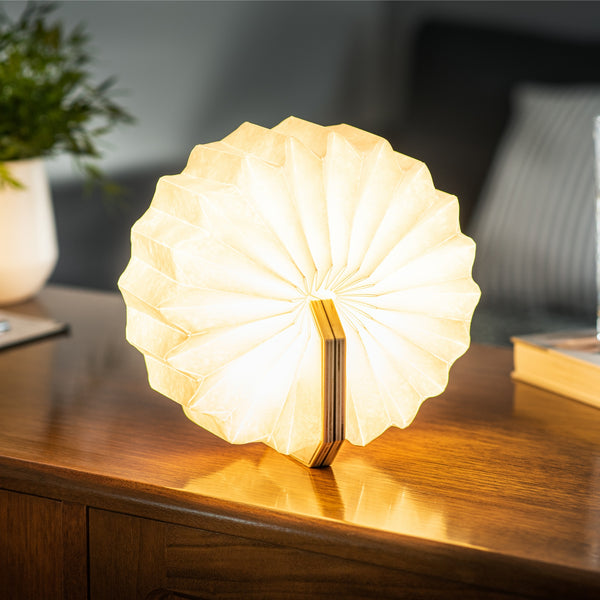Gingko Design – Lampa Smart Accordion - Naturalny Orzech, świecąca lampa w kształcie wachlarza stojąca na drewnianym meblu, przytulna atmosfera