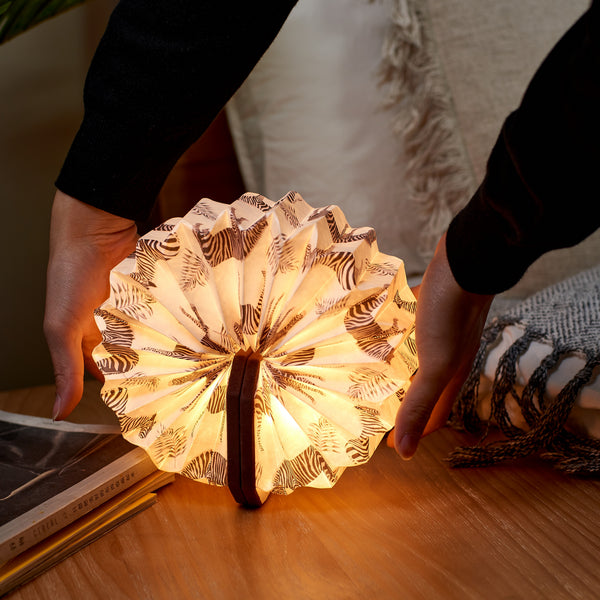Gingko Design – Lámpara Smart Accordion - Terciopelo Marrón Cebra, lámpara libro abierta con estampado de cebra, colocada sobre una mesa de madera