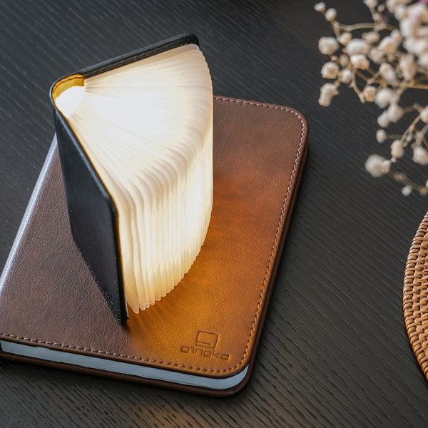 Gingko Design – Lámpara Smart Book LED - Cuero Negro - 2 Tamaños, lámpara libro abierta colocada sobre una libreta marrón