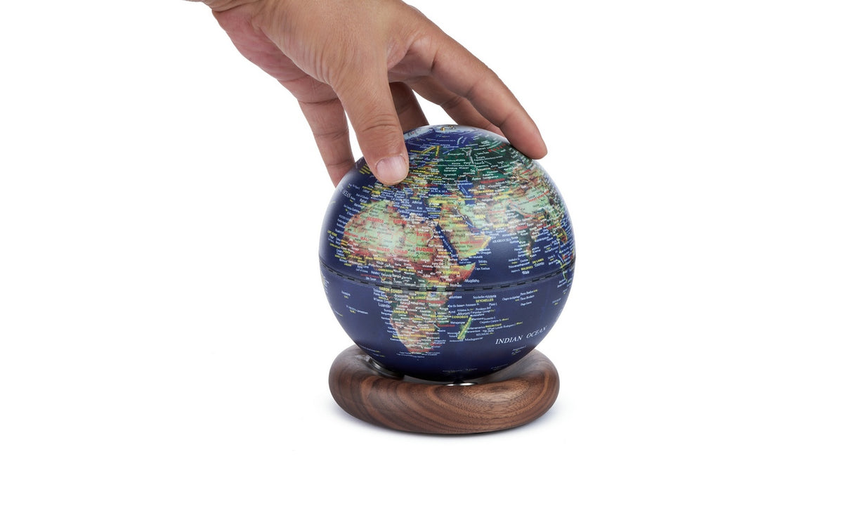 Gingko Design – Lampe Globe Atlas - Bleu Foncé / Socle Noyer - Mini, une main tourne un globe terrestre bleu sur un socle en bois