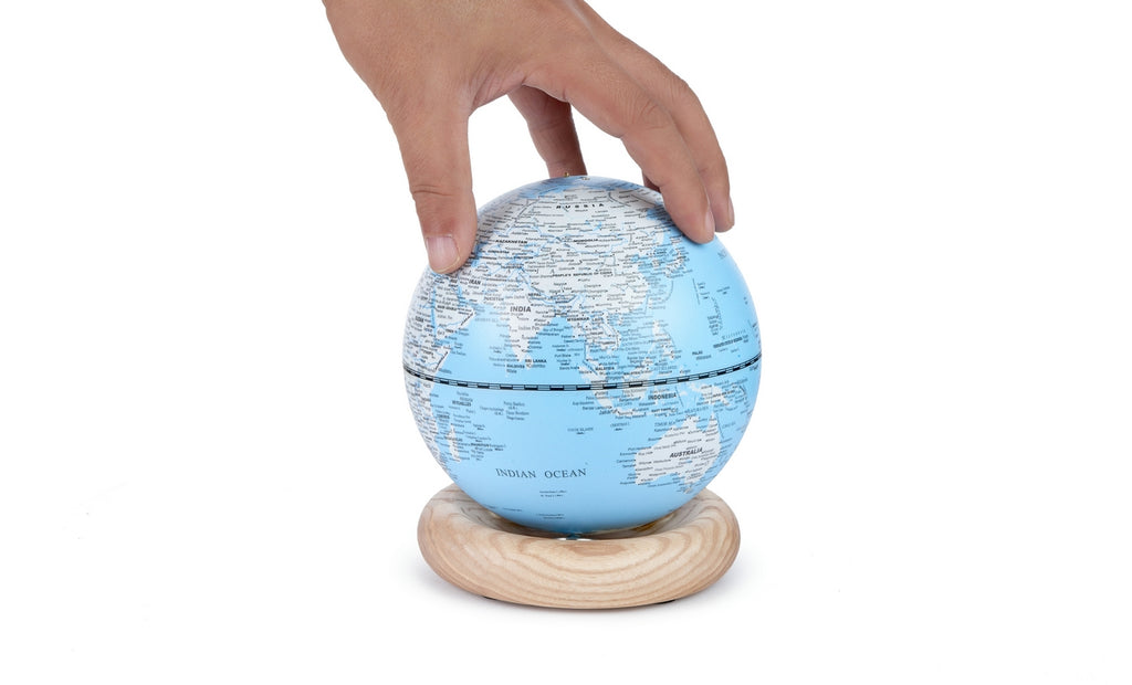 Gingko Design – Lampada Mappamondo Atlas - Blu Chiaro / Base Frassino Bianco - Mini, una mano afferra un mappamondo blu e bianco su una base in legno chiaro