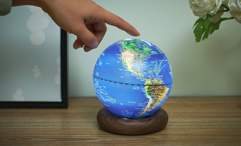 Gingko Design – Lampe Globe Atlas - Bleu Foncé / Socle Noyer - Mini, une main touche un globe terrestre lumineux posé sur un socle en bois