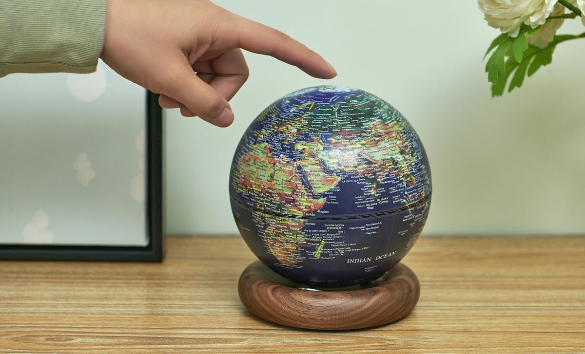 Gingko Design – Lampe Globe Atlas - Bleu Foncé / Socle Noyer - Mini, une main touche un globe terrestre posé sur un socle en bois