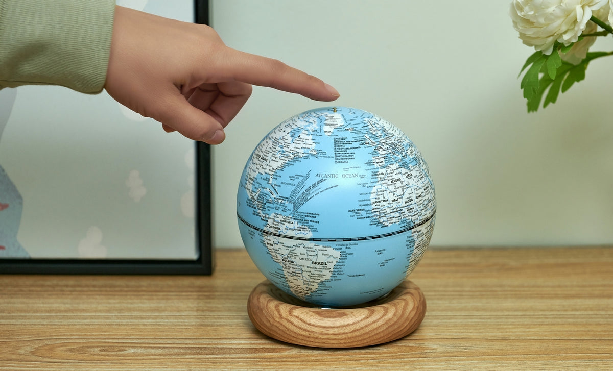 Gingko Design – Lampada Mappamondo Atlas - Blu Chiaro / Base Frassino Bianco - Mini, una mano tocca un mappamondo blu posato su una base in legno