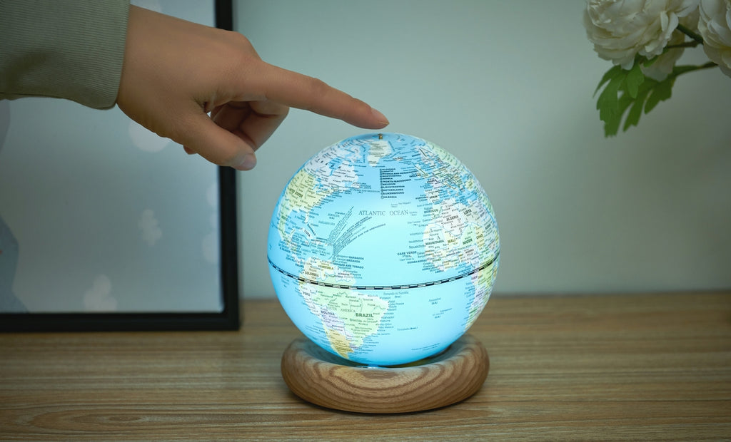 Gingko Design – Lampada Mappamondo Atlas - Blu Chiaro / Base Frassino Bianco - Mini, una mano tocca un mappamondo luminoso posato su una base in legno