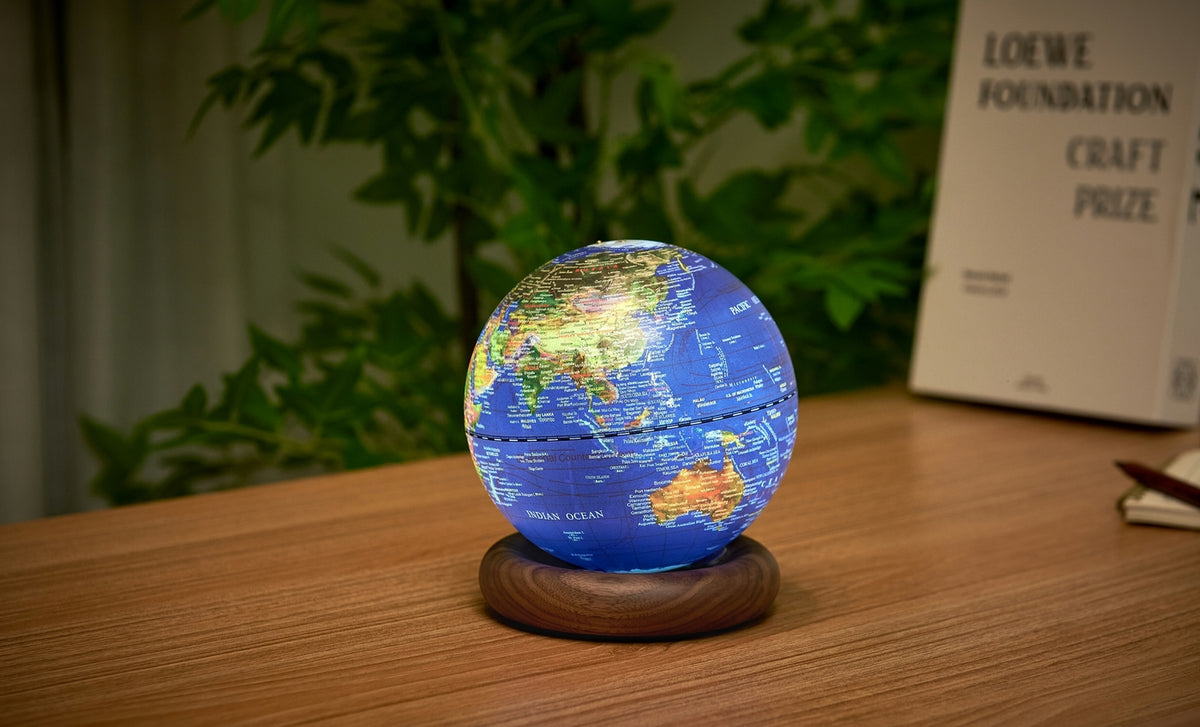 Gingko Design – Lampe Globe Atlas - Bleu Foncé / Socle Noyer - Mini, un globe terrestre lumineux posé sur un bureau en bois clair