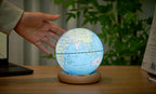 Gingko Design – Lampada Mappamondo Atlas - Blu Chiaro / Base Frassino Bianco - Mini, una mano tocca un mappamondo luminoso posato su una scrivania