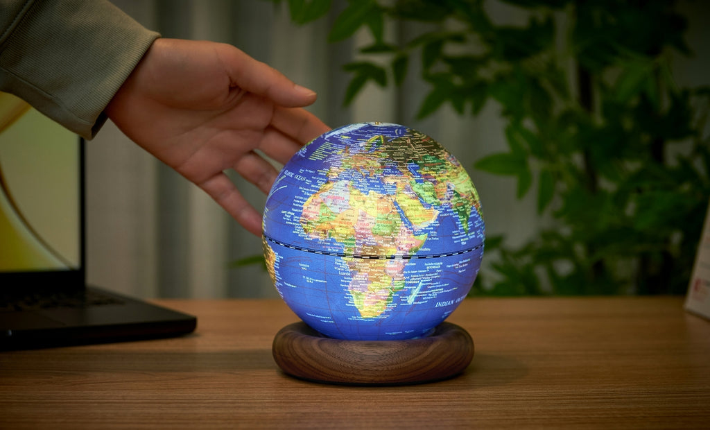 Gingko Design – Lampe Globe Atlas - Bleu Foncé / Socle Noyer - Mini, une main touche un globe terrestre lumineux posé sur un bureau