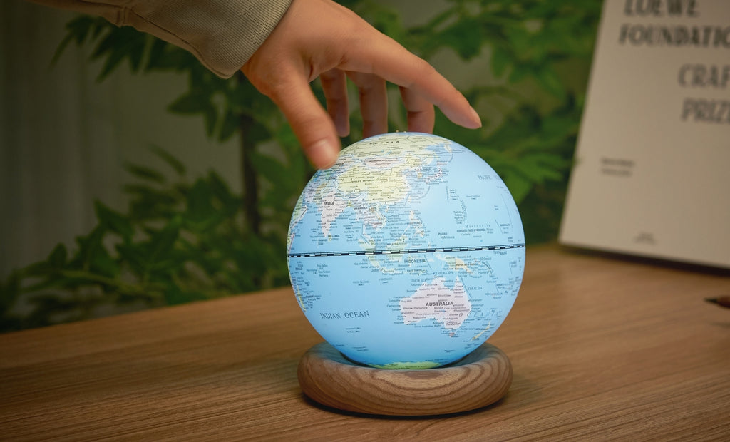 Gingko Design – Lampada Mappamondo Atlas - Blu Chiaro / Base Frassino Bianco - Mini, una mano tocca un mappamondo luminoso su una base in legno