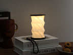 Gingko Design – Lumière Twist Hexagon - Noyer Flotté - Mini, lampe de table pliable allumée, posée sur des livres, ambiance chaleureuse
