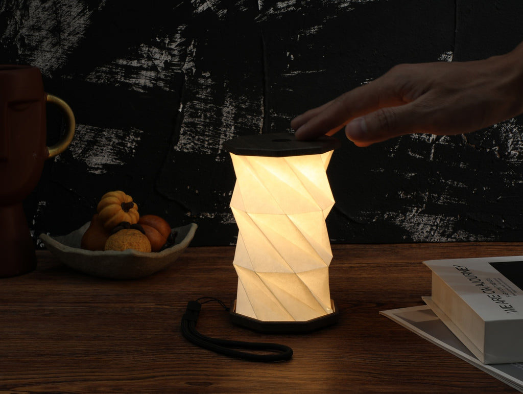 Gingko Design – Lumière Twist Hexagon - Noyer Flotté - Mini, lampe de table pliable allumée, posée sur une table en bois
