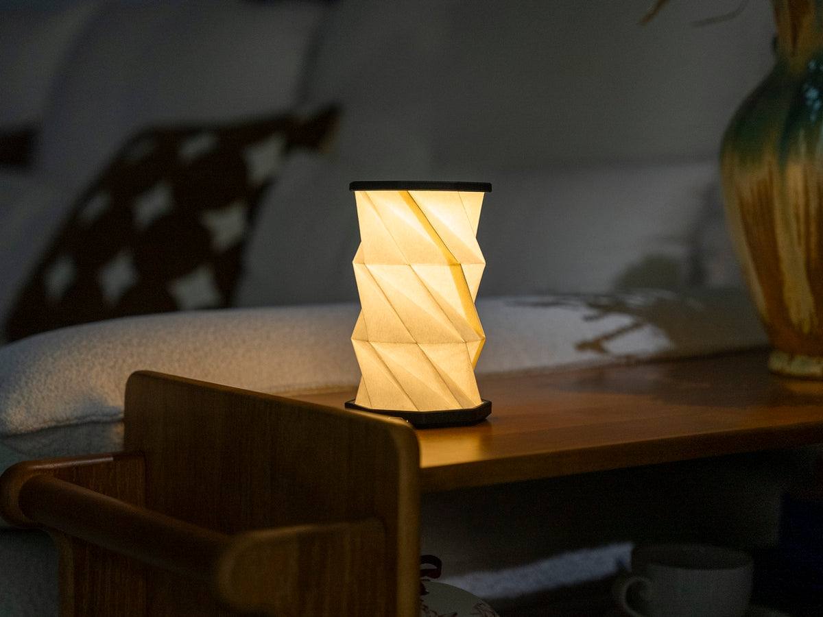 Gingko Design – Luz Twist Hexagon - Negro Pizarra - Mini, lámpara de mesa plegada de papel blanco, iluminada, sobre una mesa de madera