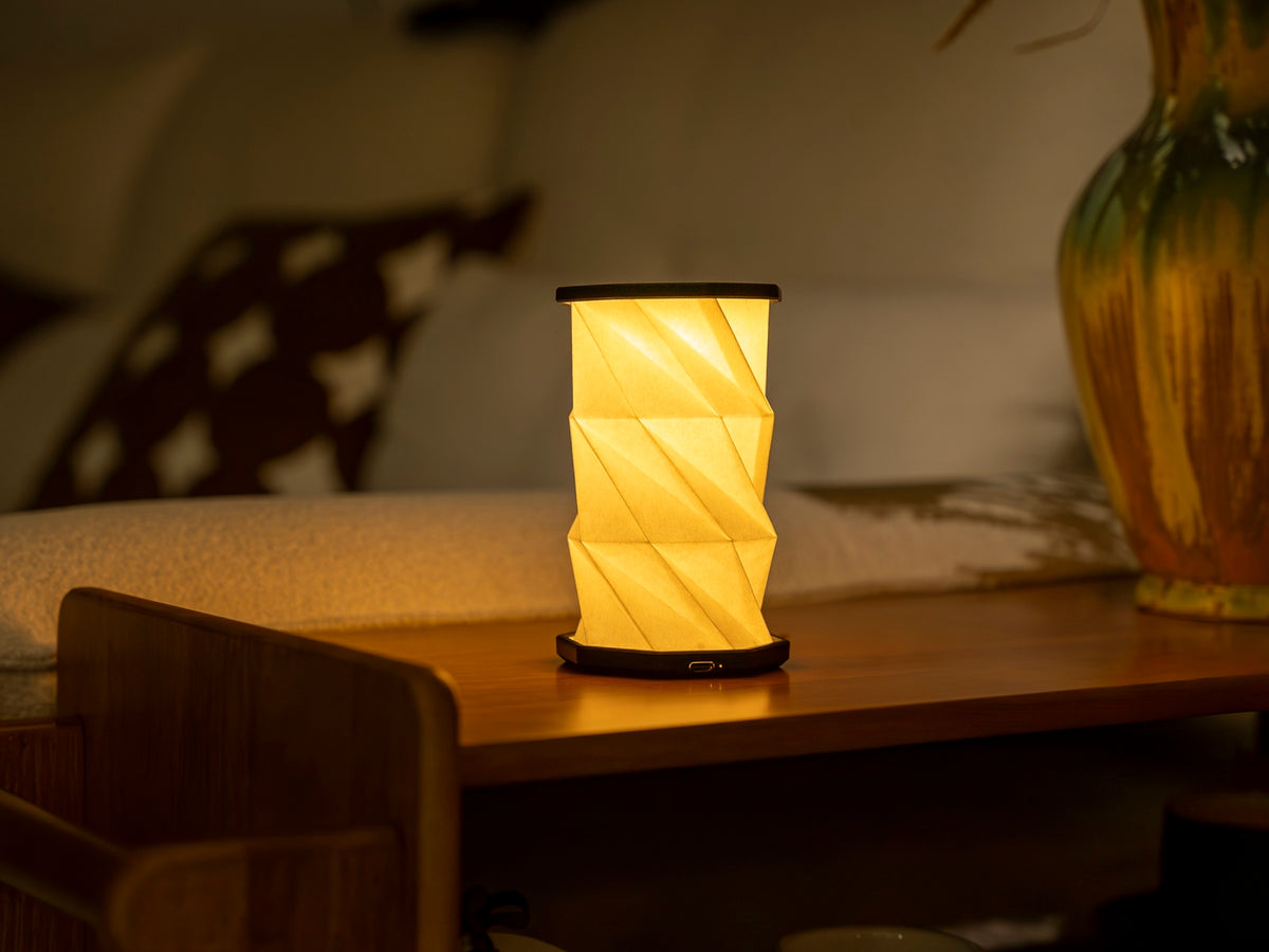 Gingko Design – Luz Twist Hexagon - Negro Pizarra - Mini, lámpara de mesa plegable encendida sobre una estantería de madera, ambiente cálido