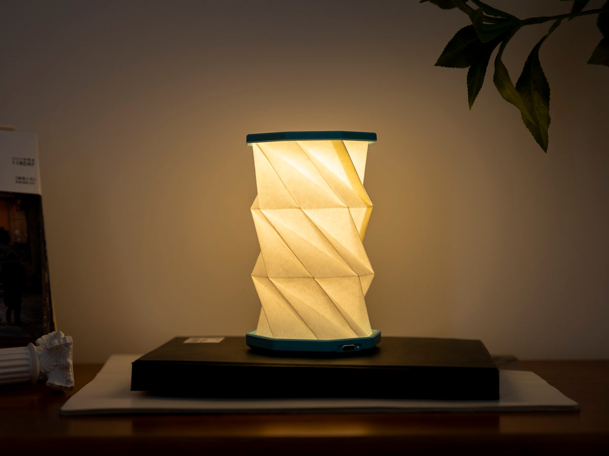 Gingko Design – Luz Twist Hexagon - Azul Laguna - Mini, lámpara de sobremesa plegable encendida, colocada sobre un libro, ambiente cálido