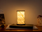 Gingko Design – Lumière Twist Hexagon - Noyer Flotté - Mini, lampe de table pliante allumée sur un bureau en bois