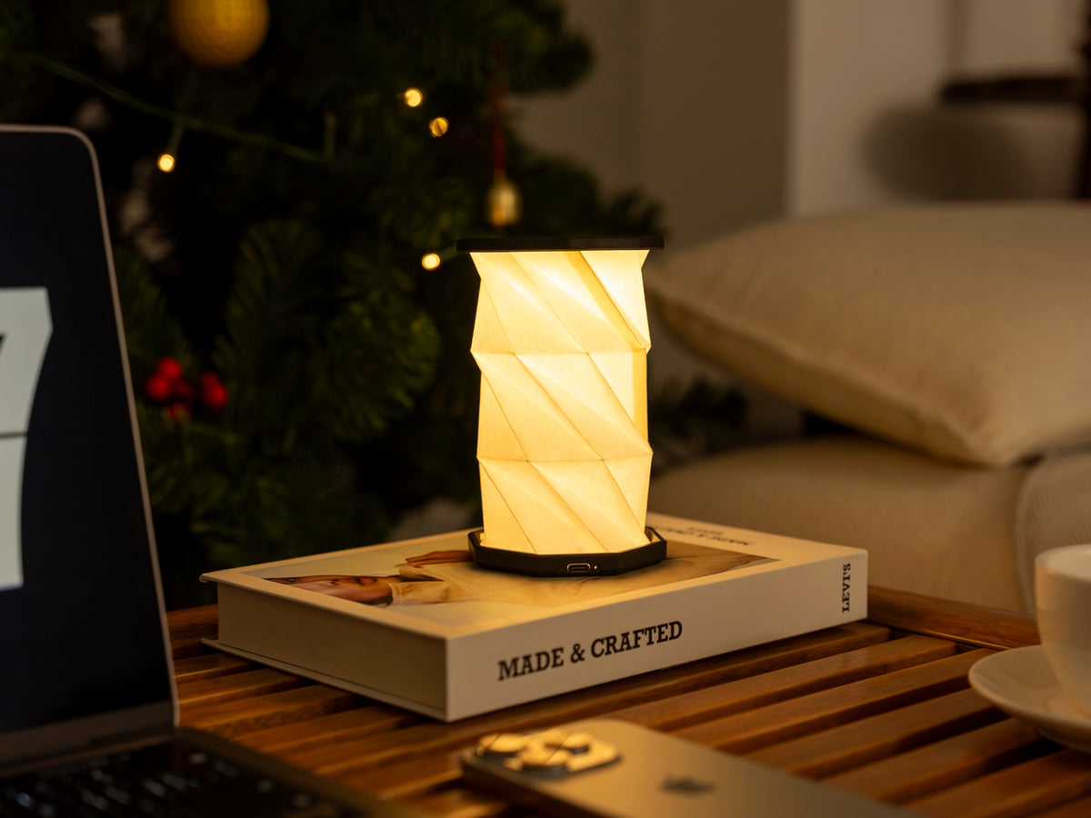 Gingko Design – Luz Twist Hexagon - Negro Pizarra - Mini, lámpara de mesa plegable encendida sobre un libro, ambiente cálido
