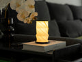 Gingko Design – Luz Twist Hexagon - Negro Pizarra - Mini, lámpara de mesa encendida sobre un libro en un salón en penumbra