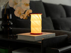 Gingko Design – Lumière Twist Hexagon - Rouge Corail - Mini, lampe de table allumée sur un livre, dans un salon sombre