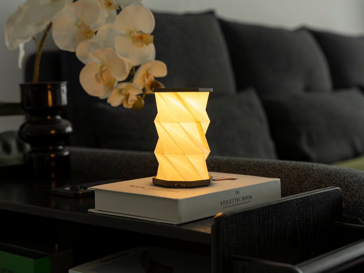 Gingko Design – Lumière Twist Hexagon - Noyer Flotté - Mini, lampe de table allumée sur un livre, dans un salon moderne