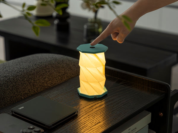 Gingko Design – Luz Twist Hexagon - Azul Laguna - Mini, lámpara de sobremesa plegable encendida, tocada por una mano, sobre una mesa de madera