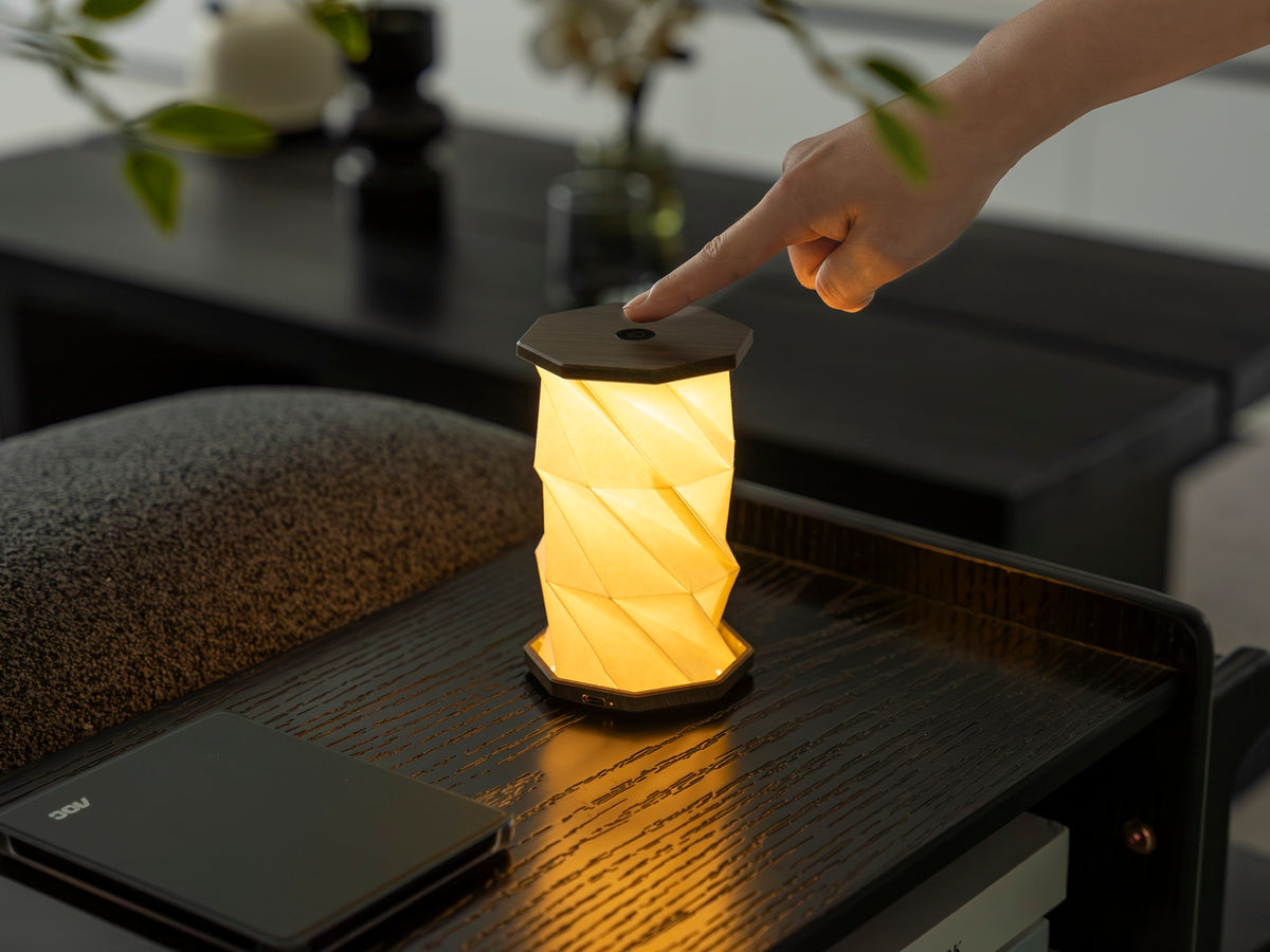 Gingko Design – Lumière Twist Hexagon - Noyer Flotté - Mini, lampe de table pliable en papier, allumée, posée sur une table en bois