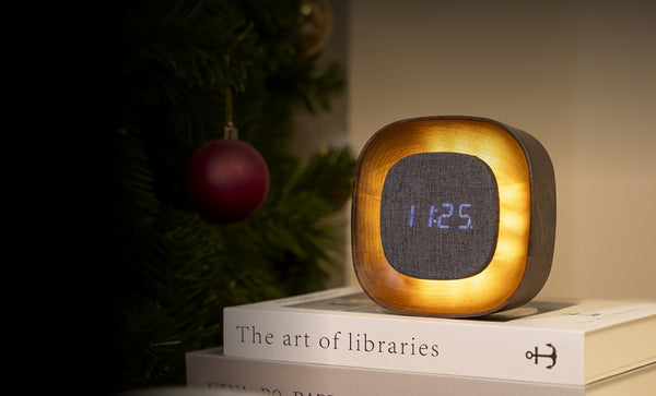 Gingko Design – Reloj Lumos - Cuadrado Nogal, despertador luminoso colocado sobre libros, cerca de un árbol de Navidad
