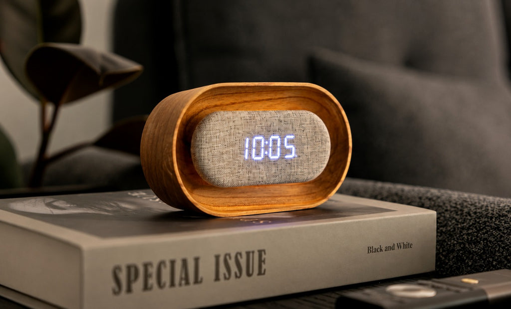 Gingko Design – Reloj Lumos - Ovalado Fresno Blanco, despertador de madera clara y tela gris sobre un libro en una mesita de noche