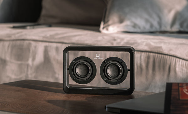 Gingko Design – Altavoz Mage Bluetooth Transparente - Madera Negra, altavoz transparente colocado sobre una mesa de madera, delante de un sofá