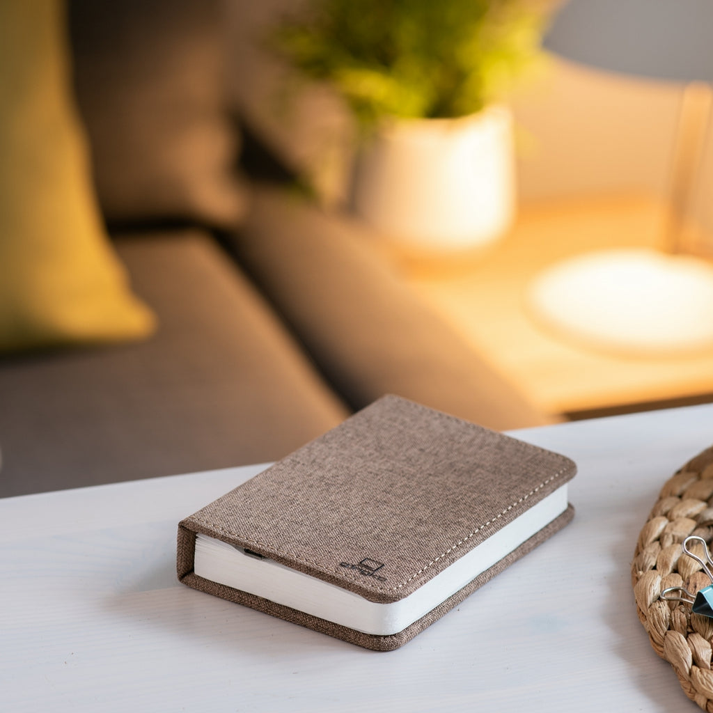 Gingko Design – Lampada Smart Book LED - Lino Marrone Caffè - Mini, un taccuino marrone posato su un tavolo bianco in un soggiorno