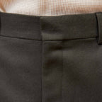 Hast – Dark Green Wool Pleated Trousers, close-up of the waistband of dark grey suit trousers, with a light-coloured jumper in the background