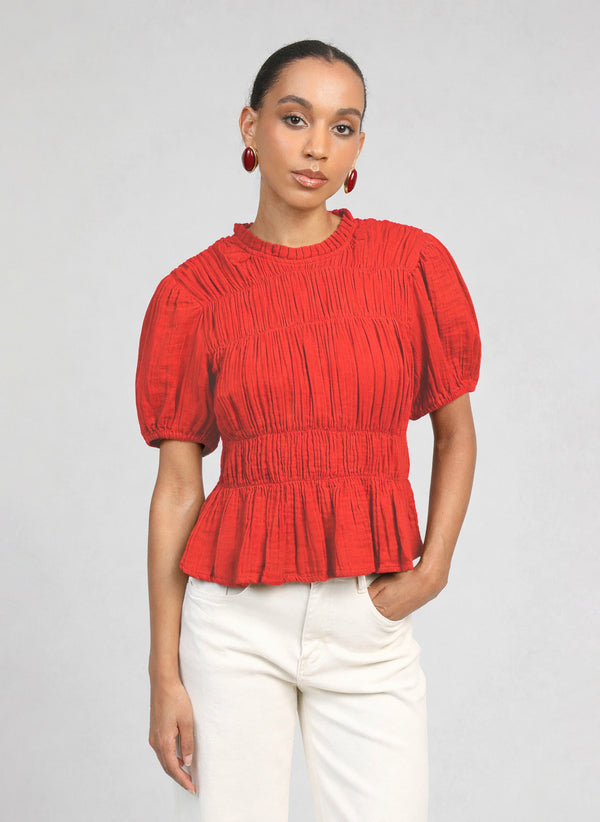 An'ge – Top Sizaya Rouge, Model trägt ein rotes plissiertes Kurzarm-Oberteil und eine helle Hose
