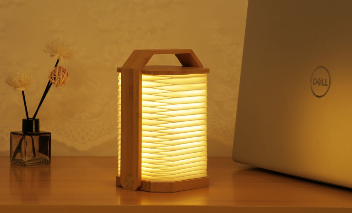 Gingko Design – Lámpara Smart Origami - Bambú Natural, lámpara plegable de madera clara, encendida, colocada sobre una mesa de madera