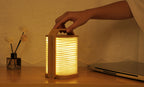 Gingko Design – Lámpara Smart Origami - Bambú Natural, una mano agarra una lámpara de madera y papel plisado encendida