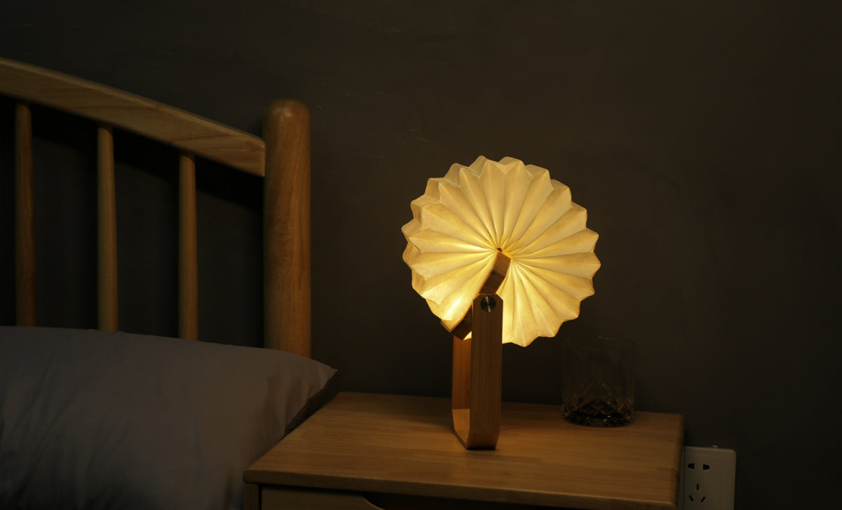 Gingko Design – Lámpara Smart Origami - Bambú Natural, lámpara de noche plegable de madera y papel, encendida, sobre una mesita de noche