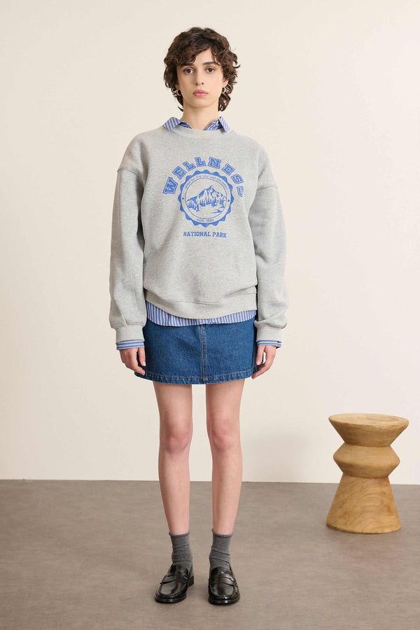Modelo mujer de frente con sudadera amplia en color gris, cuello redondo, manga larga y estampado en el pecho.