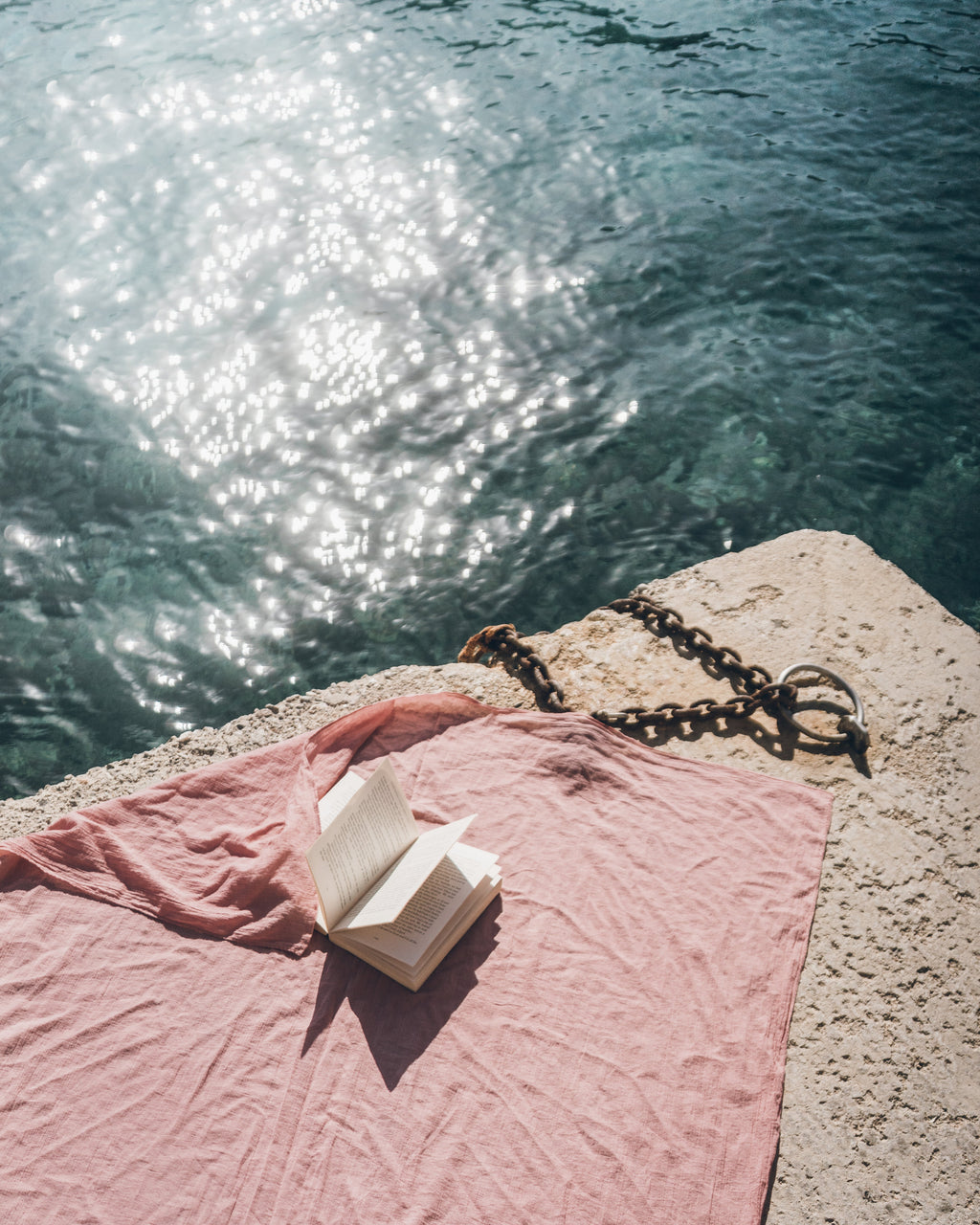 Open book on a blush pareo by the water with sunlight reflecting off the water.