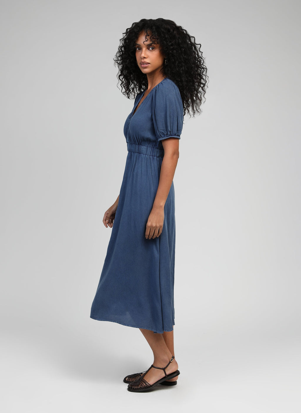 An'ge – Midikjole Omaria Indigo, kvindelig model i lang blå kjole, set i profil, lys baggrund