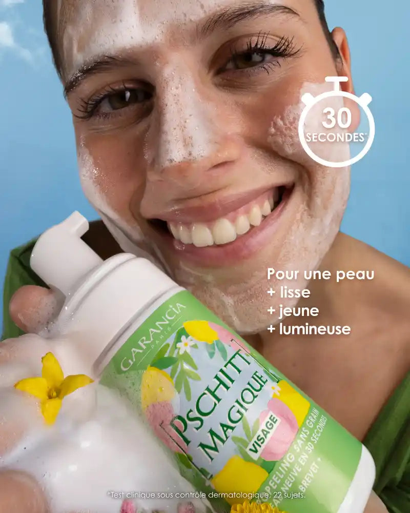 A smiling woman applies the creamy foam of Pschitt Magique Garancia to her face. She holds the limited edition bottle in front of a blue sky dotted with clouds, illustrating a moment of fresh and natural care.