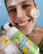 A smiling woman applies the creamy foam of Pschitt Magique Garancia to her face. She holds the limited edition bottle in front of a blue sky dotted with clouds, illustrating a moment of fresh and natural care.